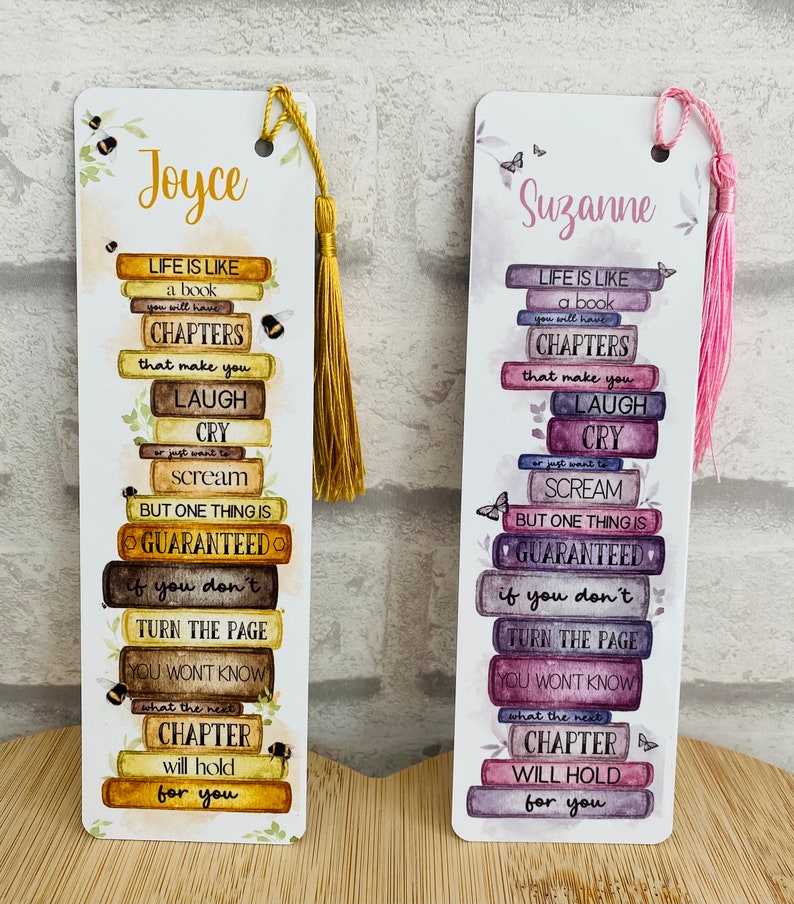 May include: Two decorative bookmarks with tassels. One bookmark has the name "Joyce" and a gold tassel, the other has the name "Suzanne" and a pink tassel. Both bookmarks feature a stack of books with inspirational text.