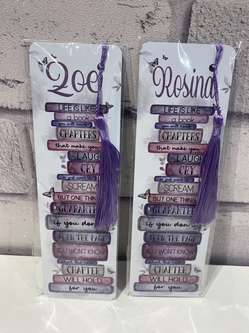 May include: Two personalized bookmarks with purple tassels. Each bookmark features a stack of books with the text "Life is like a book you will have chapters that make you laugh cry..." and a name at the top.