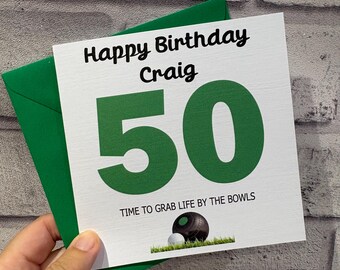 Pode incluir: Um cartão de aniversário quadrado branco com um envelope verde. O cartão apresenta o texto Happy Birthday Craig, um número 50 grande em verde, o texto TIME TO GRAB LIFE BY THE BOWLS e uma ilustração de uma bola de lawn bowls na relva.