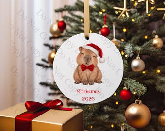 Pode incluir: Um ornamento redondo de cerâmica branca com uma capivara castanha usando um gorro de Pai Natal vermelho e uma gravata borboleta vermelha. O texto "Christmas 2026" está impresso a vermelho abaixo da capivara. O ornamento pende de uma fita dourada.