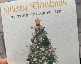 Pode incluir: Um cartão de felicitações quadrado branco com o texto dourado Merry Christmas TO THE BEST HAIRDRESSER. Abaixo do texto, há uma árvore de Natal verde decorada com enfeites cor-de-rosa e ilustrações de ferramentas de cabeleireiro.