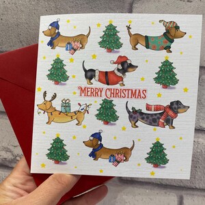 Pu&ograve; includere: Un biglietto di Natale con illustrazioni di bassotti vestiti con abiti festivi, tra cui cappelli e maglioni, con regali e alberi di Natale. Le parole "MERRY CHRISTMAS" sono stampate in rosso. Il biglietto &egrave; tenuto con una busta rossa.