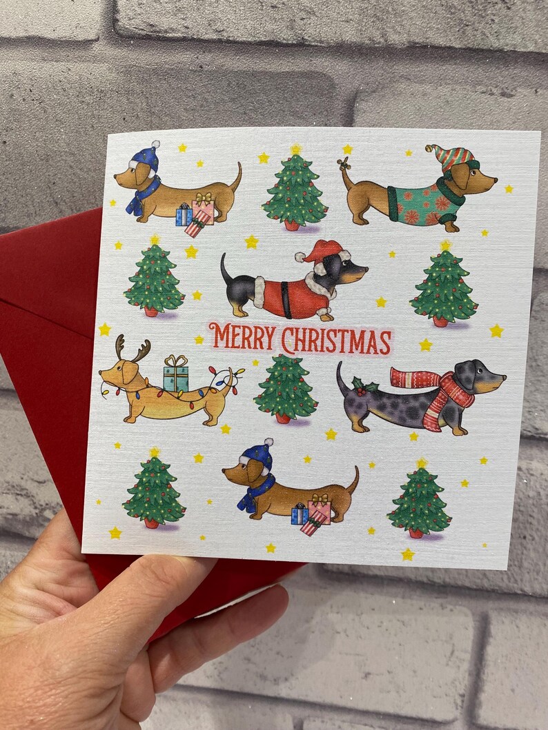 Pu&ograve; includere: Un biglietto di Natale con illustrazioni di bassotti vestiti con abiti festivi, tra cui cappelli e maglioni, con regali e alberi di Natale. Le parole "MERRY CHRISTMAS" sono stampate in rosso. Il biglietto &egrave; tenuto con una busta rossa.