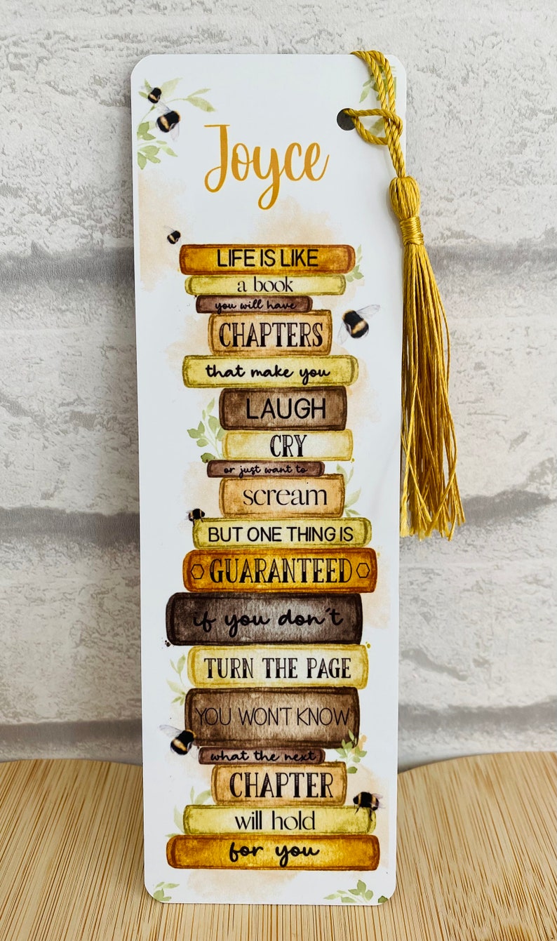 May include: White bookmark with a gold tassel, featuring a stack of book illustrations and the text "Life is like a book..." with the name "Joyce" at the top. Bumblebee and leaf accents decorate the design.