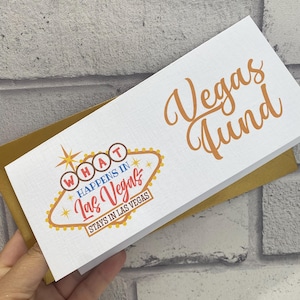 Può includere: Una busta rettangolare bianca con interno color oro. La parte anteriore presenta la scritta "Vegas Fund" in corsivo arancione e la grafica di un cartello giallo e arancione con il testo "WHAT HAPPENS IN Las Vegas STAYS IN LAS VEGAS".