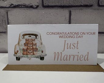 Pode incluir: Um cartão de casamento com uma ilustração de um carro vintage transportando malas empilhadas. O cartão diz "Parabéns pelo seu dia de casamento" e "Just Married" em uma fonte suave e elegante. O cartão está em uma base marrom.