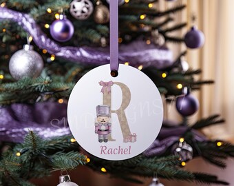 Pode incluir: Um enfeite de Natal redondo e branco pendurado por uma fita lavanda. O enfeite apresenta uma letra R grande dourada decorada com flocos de neve, um laço rosa, um boneco quebra-nozes rosa, pequenos presentes e o nome Rachel em rosa.