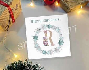Pode incluir: Um cartão de felicitações quadrado branco com o texto Merry Christmas em azul-petróleo. No centro, uma letra R grande decorada com um quebra-nozes rosa e uma guirlanda de flocos de neve azul-claros e prateados sobre um fundo branco.