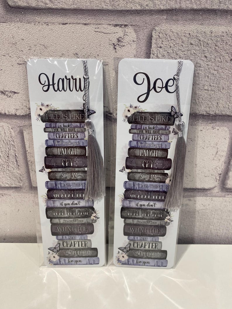May include: Two white bookmarks with gray tassels. Each bookmark features a stack of books with the words "LIFE IS LIKE" and other phrases. One bookmark says "Harry" and the other says "Joe".