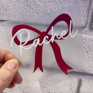 May include: A white vinyl decal with the name "Rachel" in a white cursive font on a burgundy bow.
