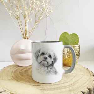 Può includere: Una tazza in ceramica bianca con un bordo e un manico grigi presenta un disegno a matita in bianco e nero di un piccolo cane bianco con pelo soffice. La tazza è appoggiata su una superficie di legno.