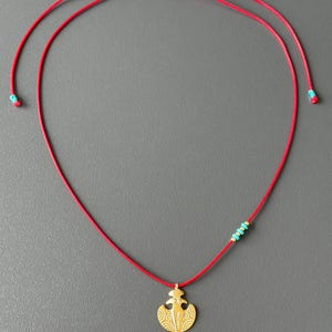 May include: A red cord necklace with a gold pendant featuring a stylized design. The pendant is attached to the cord with a small turquoise bead.