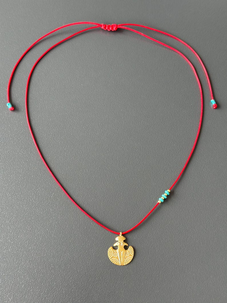 May include: A red cord necklace with a gold pendant featuring a stylized design. The pendant is attached to the cord with a small turquoise bead.