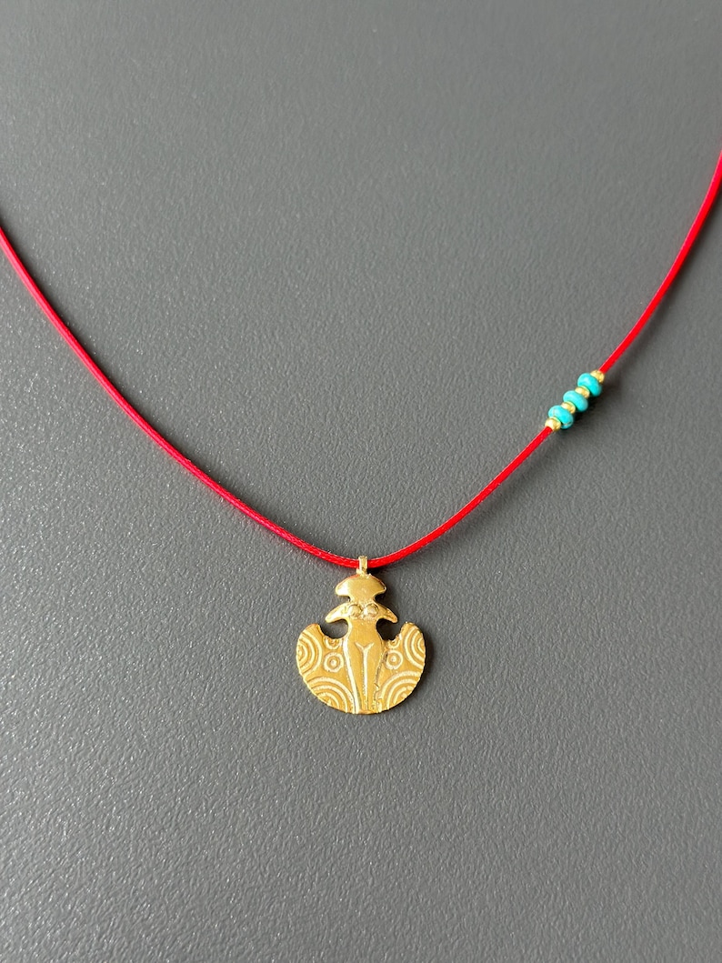 May include: A gold pendant necklace with a stylized female figure design. The necklace is on a red cord with turquoise beads.