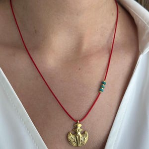 May include: A gold pendant necklace with a red cord and turquoise beads. The pendant features a stylized female figure with wings.