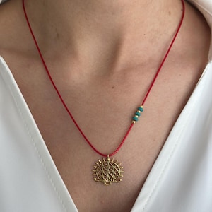 May include: A gold pendant necklace with a red cord. The pendant has a detailed, sun-like design. The necklace also features small turquoise beads. The necklace is worn against a white shirt.