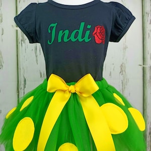 May include: A green tulle tutu skirt with yellow polka dots and a yellow satin bow. The skirt is paired with a black t-shirt with the name "Indi" and a red glitter rose.