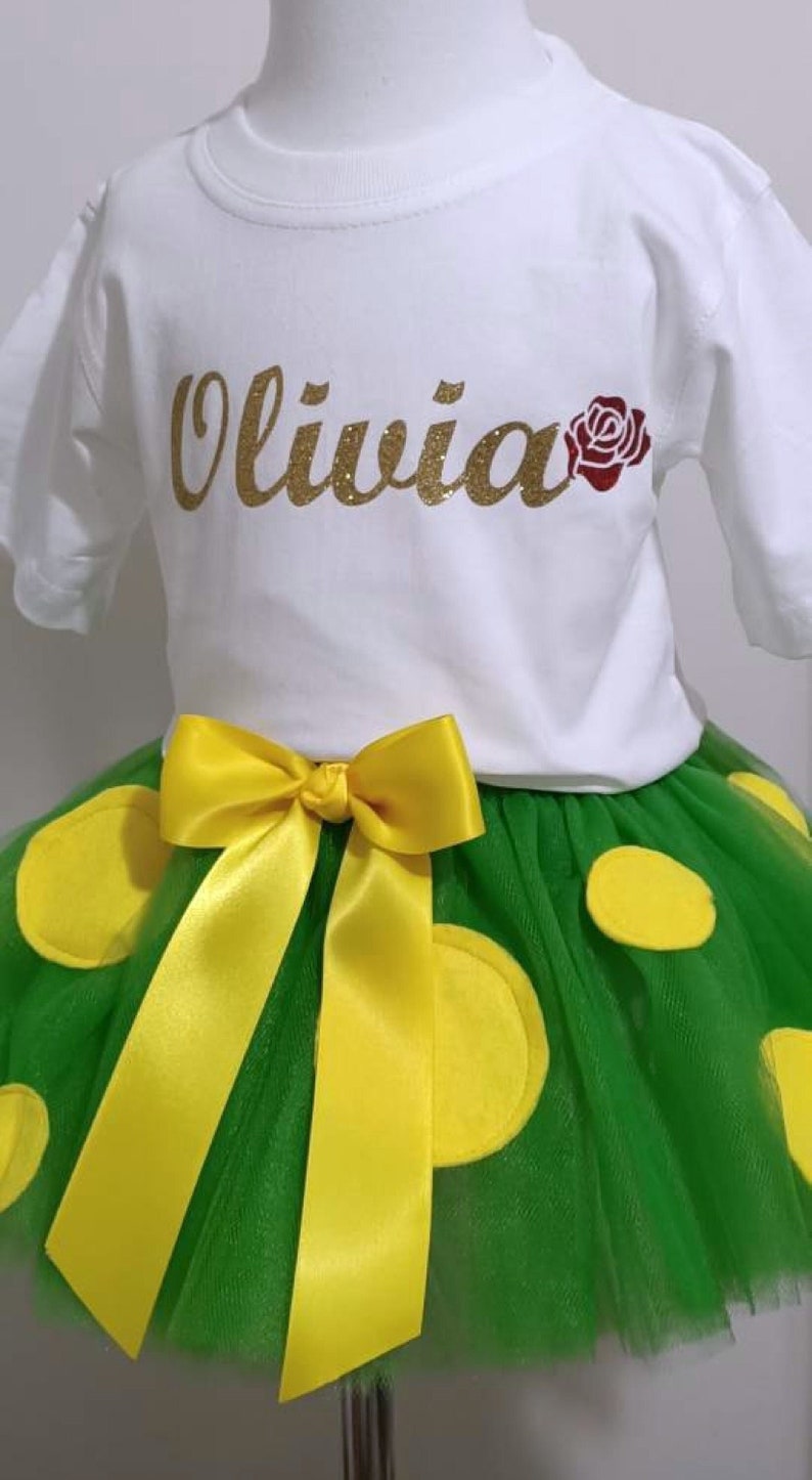 May include: A white t-shirt with the name "Olivia" in gold glitter lettering and a red rose. The shirt is paired with a green tulle skirt with yellow polka dots and a yellow satin bow.