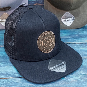 May include: Three black, grey, and green mesh baseball caps with a black leather patch on the front of the black cap that says "Original Est. 1994" and a white label on the side of each cap that says "Snapback".