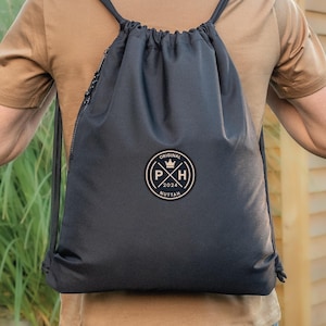 Peut inclure: Sac à dos noir avec cordon de serrage, orné d'un écusson circulaire avec le texte "ORIGINAL P X H 2024 NUTTAH" et une couronne. Le sac a des cordons noirs et une fermeture éclair. La personne porte un t-shirt marron.
