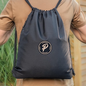 May include: A black drawstring backpack with a white and black circular logo. The backpack has black shoulder straps and a side zip. The person is wearing a brown t-shirt.