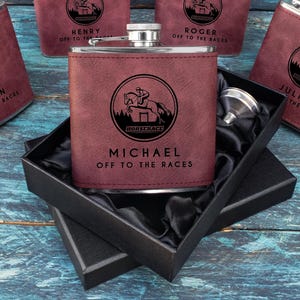May include: A collection of maroon-colored flasks with a stainless steel top. Each flask features a circular graphic of a horse and rider, with names and the phrase "Off to the Races" engraved below. One flask is in a black gift box.