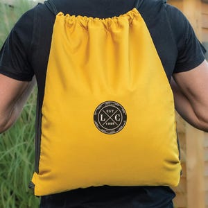 May include: A bright yellow drawstring backpack with black drawstrings. The backpack features a circular logo with the text "EST 1988" and the letters "LXC". The person is wearing a black t-shirt and blue jeans.