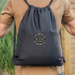 Puede incluir: Mochila negra con cordón y logotipo circular blanco con las letras "LXC" y el año "1998". La mochila tiene cordones negros y una cremallera. La persona lleva una camiseta marrón.