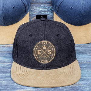 May include: Three baseball caps with a two-tone design. The caps have a blue crown and a tan brim. One cap has a circular patch with the text "COD EXS 1993 KING". The caps are on a blue wooden surface.