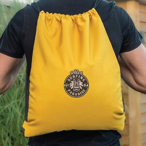 May include: A bright yellow drawstring backpack with a black logo that reads "Alex Surfing Paradise" in a circular design. The backpack is worn over the shoulders, with the drawstring closure visible at the top.