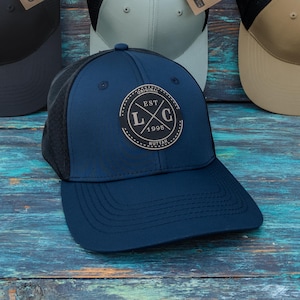 May include: Four baseball caps with mesh backs and solid front panels in black, light grey, tan, and navy blue. The navy blue cap features a circular patch with the text EST 1995 L C ORIGINAL RUTTAH. Each cap includes a brown cardboard hang tag.