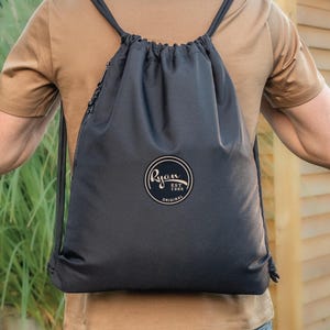 May include: Black drawstring backpack with a circular logo reading "Ryan Est. 1988 Original". The bag has black drawstrings and a zipper. The person is wearing a brown t-shirt.