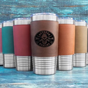May include: An assortment of insulated tumblers in shades of pink, teal, burgundy, brown, tan, and grey. Each tumbler has a stainless steel base and a leather sleeve. The brown tumbler features the text "Outdoor Hunting" with a skull and crosshair design.