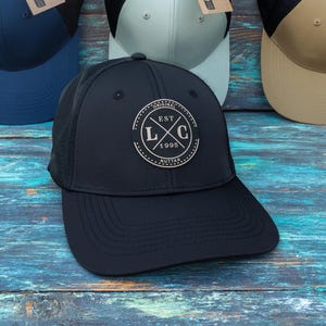 May include: Four baseball caps with mesh backs and solid front panels in navy blue, light grey, tan, and black. The black cap features a circular patch with the text EST L C 1995. Each cap includes a brown cardboard tag stating made from 100% recycled fabric.