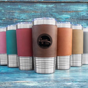 May include: An assortment of insulated tumblers in shades of pink, teal, burgundy, brown, tan, and grey. Each tumbler has a stainless steel base and a leather sleeve with the text "Ryan EST 1986 ORIGINAL".