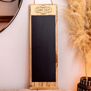 Può includere: Una lavagna con cornice in legno con un cartello che dice "Games Night" appesa a un muro bianco. La lavagna è nera e ha un gancio in corda.
