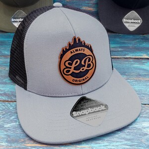 May include: A gray and black mesh trucker hat with a brown leather patch that says "Always Original" and has a stylised "LB" logo. The hat has a black mesh back and a gray front with a black Snapback label.
