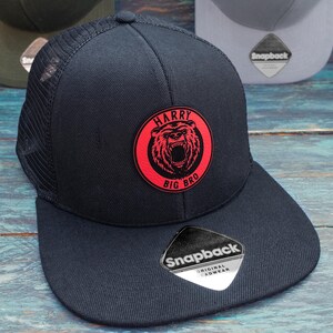 May include: Black mesh baseball cap with a red circle patch featuring a bear with the text "HARRY BIG BRO". The cap has a black adjustable strap with a white "Snapback Original Headwear" label.