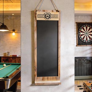 Pode incluir: Um quadro-negro retangular com moldura de madeira e corda para pendurar. A seção superior apresenta o texto "RYAN'S SCOREBOARD" e um brasão. O quadro-negro está pendurado em uma parede de concreto, com uma mesa de bilhar e um alvo de dardos visíveis ao fundo.