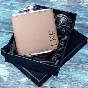 Könnte beinhalten: Eine braune, lederbezogene Edelstahl-Taschenflasche mit einem silbernen Verschluss und den eingravierten Initialen "LKP" auf der Vorderseite. Die Flasche befindet sich in einer schwarzen Box mit schwarzem Satin-Futter.