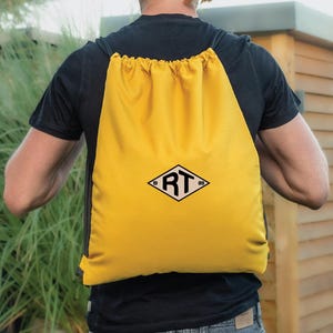 May include: A yellow drawstring backpack with black drawstrings and a black and white logo. The logo features the letters "RT" inside a diamond shape, with the numbers "19" and "89" on either side. The backpack is made of a smooth material, possibly nylon.