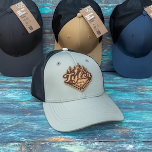 May include: Four baseball caps with mesh backs and solid front panels in black, tan, navy blue, and light grey. The light grey cap features a diamond-shaped wooden patch with a forest graphic and the text Tyler Since 83 Outdoor Adventure.
