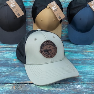 May include: Four baseball caps with mesh backs and solid front panels in black, tan, navy blue, and light grey. The light grey cap features a circular brown patch with a fish illustration and the text HARRY EST RATHER BE FISHING.