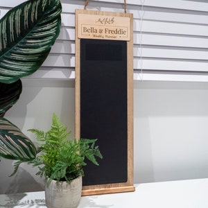 Pode incluir: Uma lousa enquadrada em madeira com uma placa que diz "Bella & Freddie Weekly Planner". A lousa é preta e tem uma pequena prateleira de madeira na parte inferior.