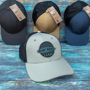 May include: Four baseball caps with mesh backs and solid front panels in black, tan, navy blue, and light grey. The light grey cap features a circular patch with vertical stripes and the text SURF RIDER LIAM. Each cap includes a brown paper tag.