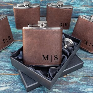May include: Five brown leather-covered hip flasks with silver tops, each engraved with initials. One flask is open in a black box with a funnel. The flasks are on a blue wooden surface. The initials are T|S, D|H, X|F, and M|S.