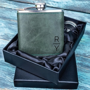 Könnte beinhalten: Eine grüne, lederbezogene Flachmann mit silbernem Verschluss, präsentiert in einer schwarzen Geschenkbox. Der Flachmann trägt die Initialen "R" und "Y". Die Box ist mit schwarzem Satin ausgekleidet.