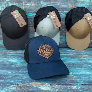 May include: Four baseball caps with mesh backs and solid front panels in black, light grey, tan, and navy blue. The navy blue cap features a brown diamond-shaped patch with a forest graphic and the text Tyler Since 83 Outdoor Adventure.