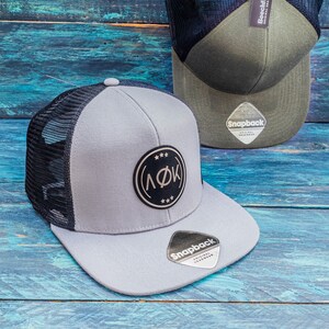 Puede incluir: Una gorra de camionero de malla gris y negra con un parche circular blanco y negro que dice "(ΛΦΚ)" y tres estrellas. La gorra tiene una parte trasera de malla negra y una parte delantera gris con una etiqueta "Snapback Original Headwear".