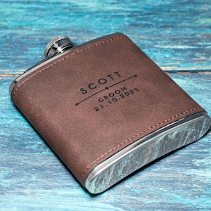 May include: A brown leather-covered flask with a silver cap and trim. The flask is personalized with the name "SCOTT", the word "GROOM", and the date "21.10.2025" in black lettering. The flask is set against a blue wooden background.